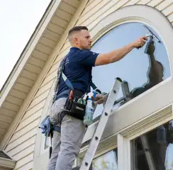 Professional window cleaner on a ladder using a squeegee to clean an exterior arched window, Austin, TX.